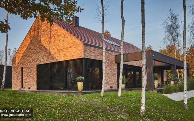 Indywidualny projekt domu z elewacją z cegły z żaluzjami zewnętrznymi dużymi oknami antracytowymi i ciekawym pomysłem na drewniany taras z dekoracyjną ramą - zdjęcia z realizacji